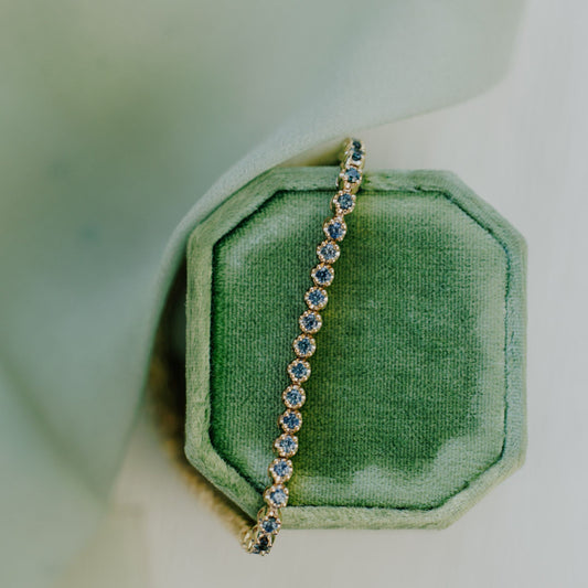 a close up shot of a teal sapphire tennis bracelet set on a green velvet jewelry box with a soft white background.