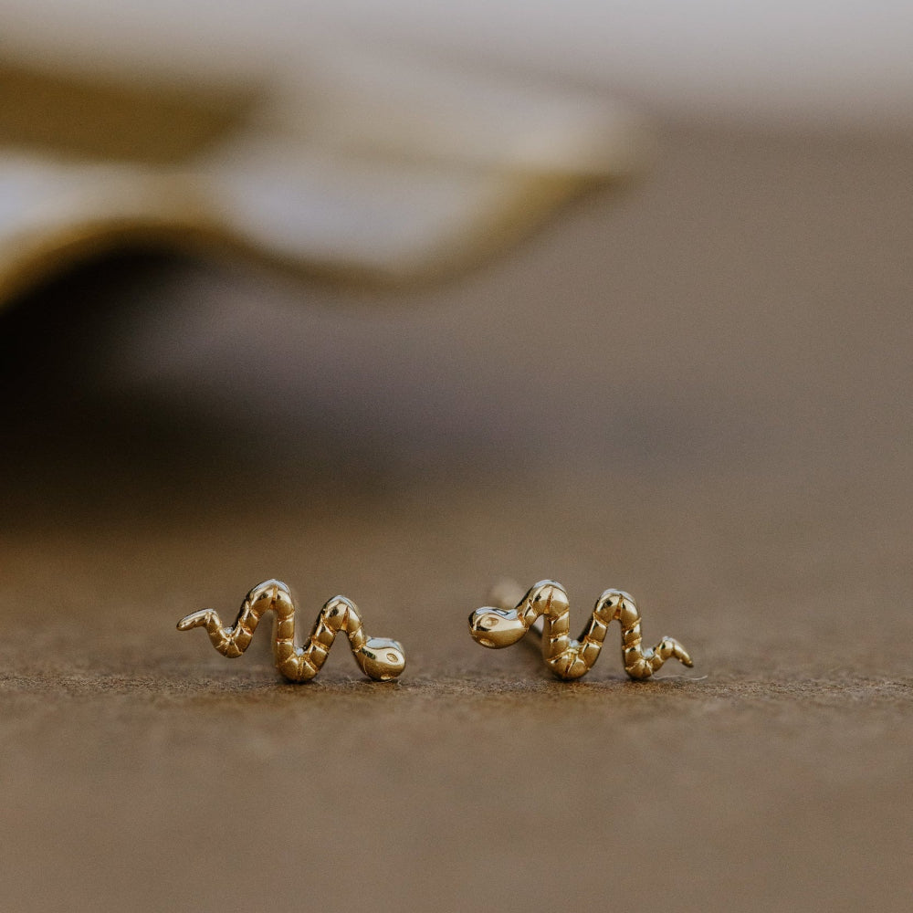 Gold snake-shaped earrings on a beige surface with a blurred background.