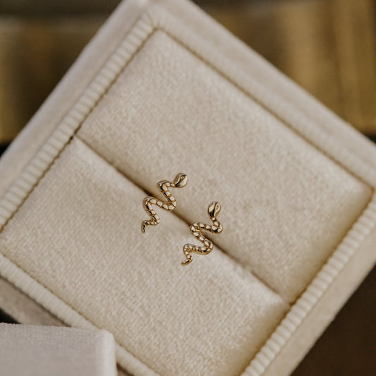 Gold snake-shaped earrings on a beige jewelry box.