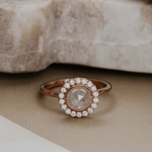 A rose gold halo ring featuring a central round rose-cut diamond surrounded by a halo of smaller clear stones, resting on a neutral beige surface with a blurred stone in the background.