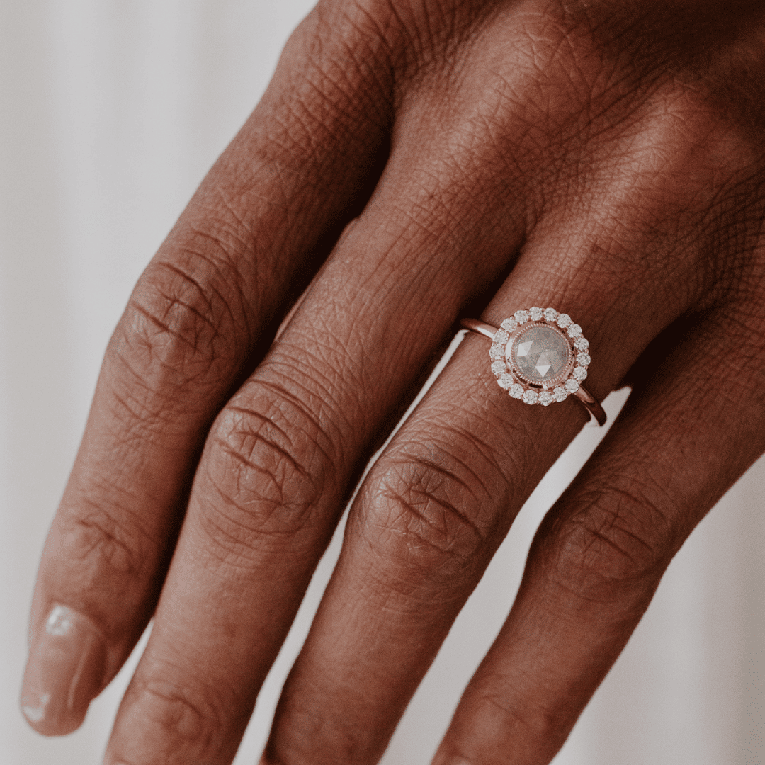 A close-up of a hand wearing a rose gold ring with a round clear center stone and a halo of small clear stones.