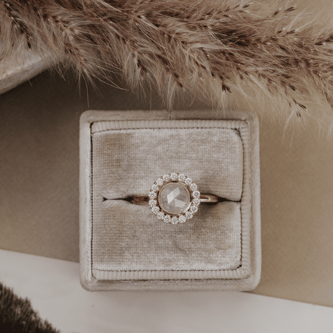 A rose gold ring with a central round-cut clear gemstone surrounded by a halo of small clear stones, displayed in a square beige velvet ring box. The box is styled with dried pampas grass on a neutral beige background.