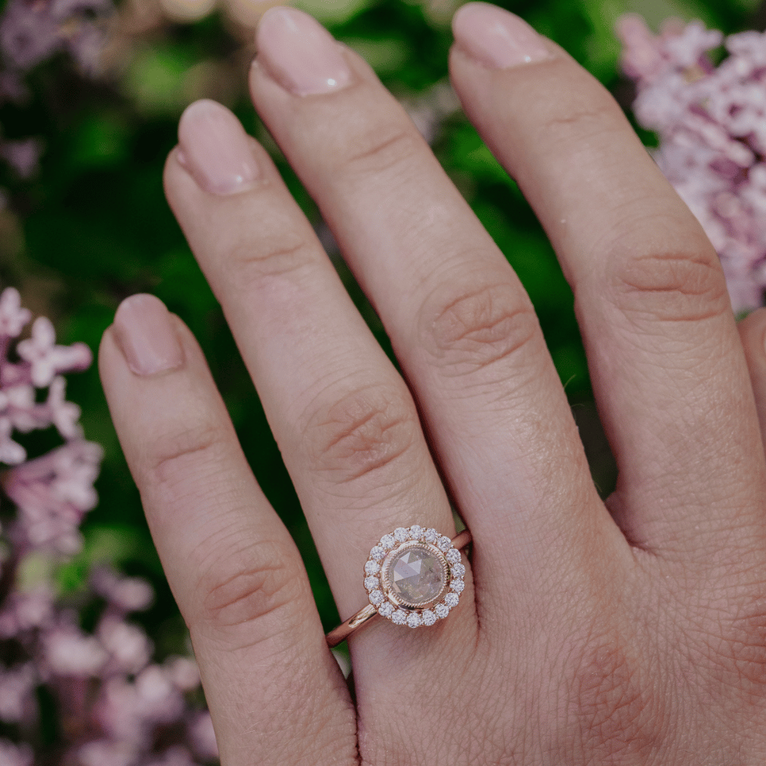 A close-up of a hand wearing a rose gold halo ring with a central round moonstone and small clear accent stones, set against a soft-focus background of green foliage and pink flowers.