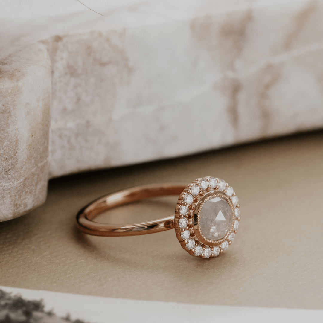 A rose gold ring with a round halo of small clear stones and a central diamond-cut stone, resting on a textured beige surface with a blurred stone prop in the background.