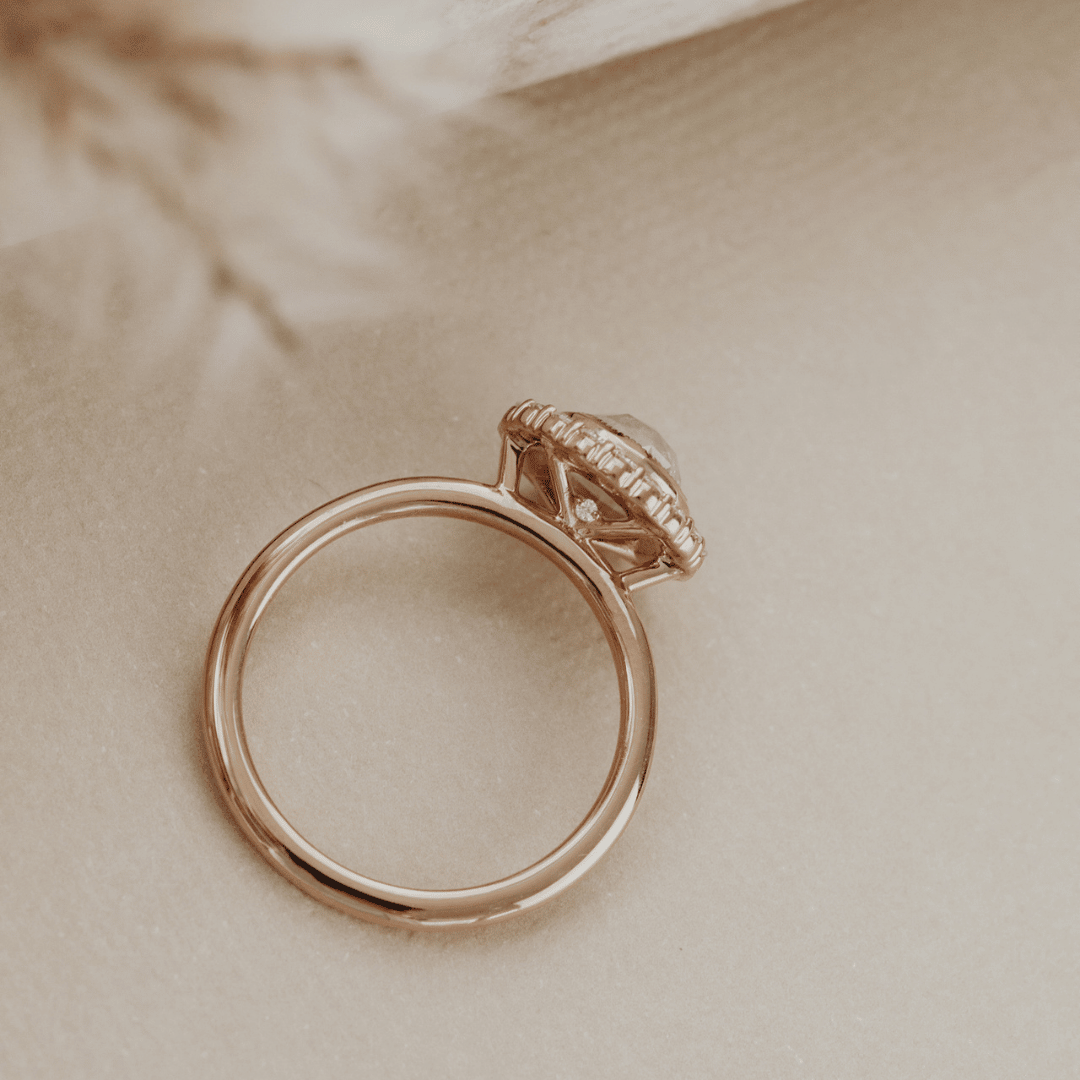 A rose gold ring with a textured pavé setting and a clear center stone, resting on a soft beige textured surface.