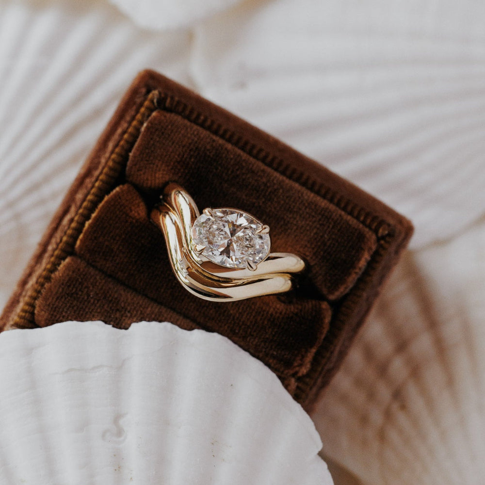 The Capri Oval Ring in 14k yellow gold featuring a lab-grown oval diamond prong set on a double wavy band, displayed in a brown velvet ring box.
