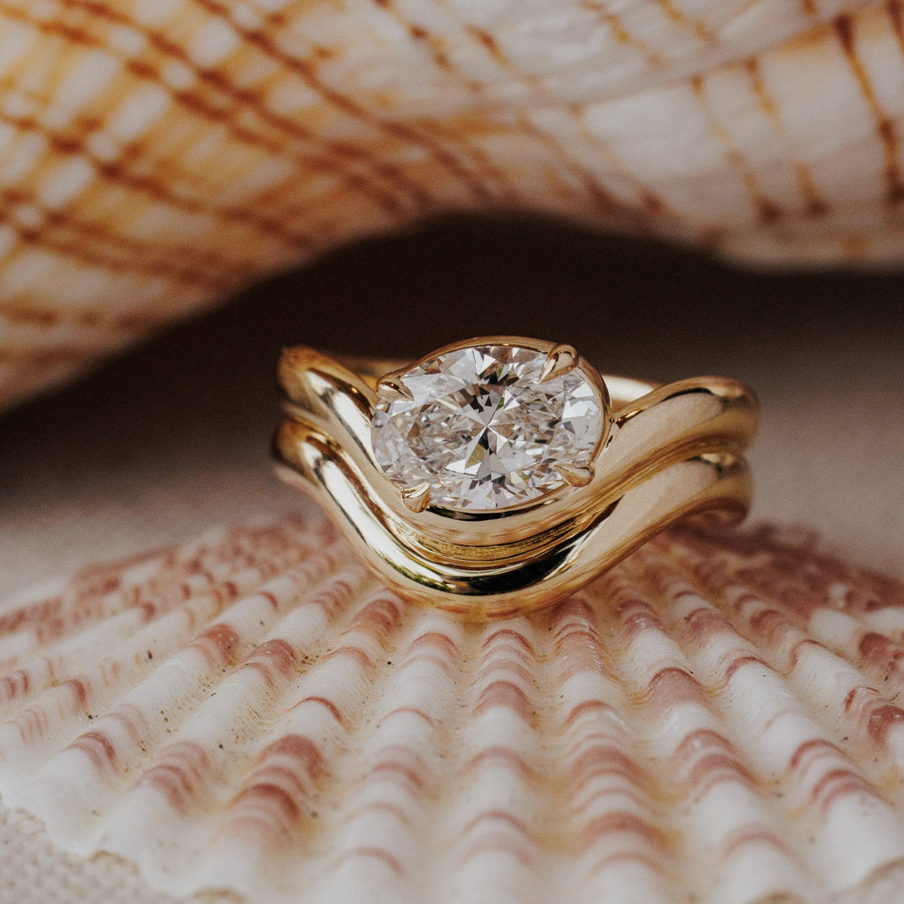 The Capri Oval Ring in 14k yellow gold featuring a lab-grown oval diamond prong set on a double wavy band, displayed inside a seashell. This 14k gold ring features an oval diamond center stone set in prongs on a double wavy band. This ring is made-to-order based on the selected customizations.