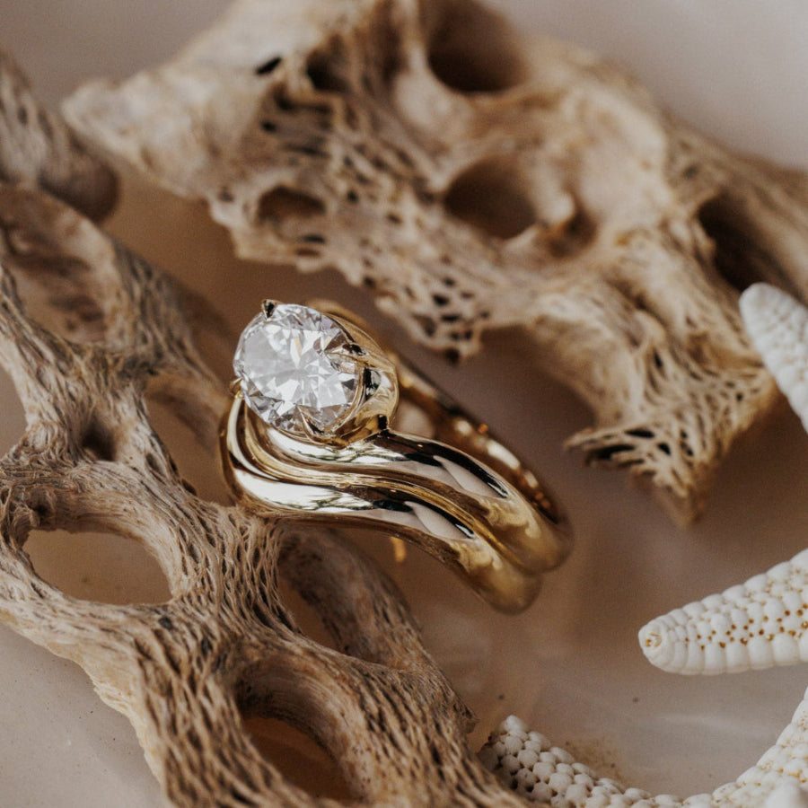 The Capri Oval Ring in 14k yellow gold featuring a lab-grown oval diamond prong set on a double wavy band, resting on coral with a starfish in the background.