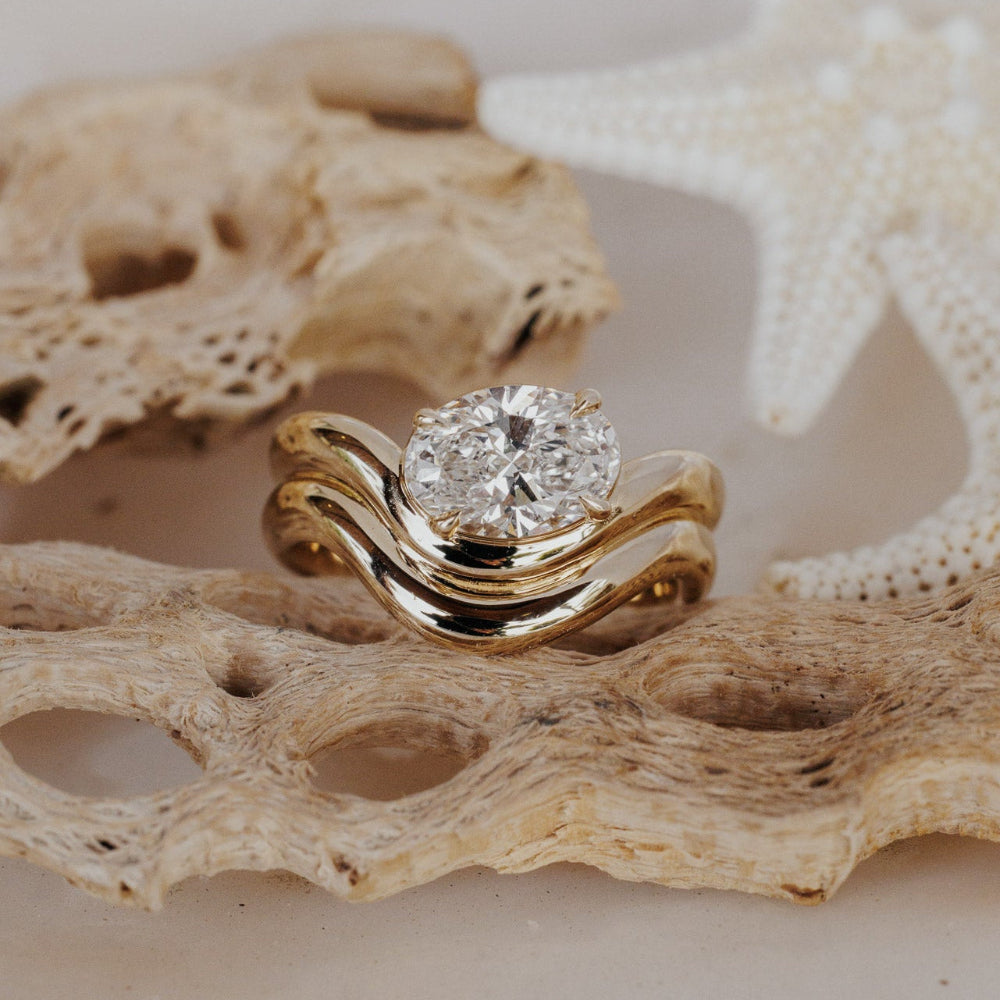 The Capri Oval Ring in 14k yellow gold featuring a lab-grown oval diamond prong set on a double wavy band, displayed on coral. This 14k gold ring features an oval diamond center stone set in prongs on a double wavy band. This ring is made-to-order based on the selected customizations.