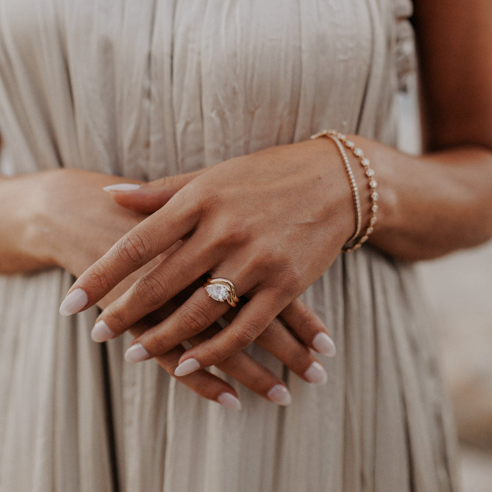 The Capri Pear Ring in 14k yellow gold featuring a lab-grown pear diamond prong set on a double wavy band, worn on the ring finger alongside stacked diamond bracelets.
