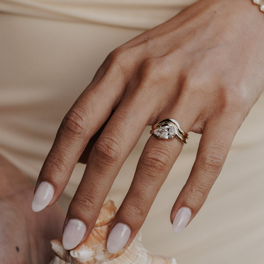 The Capri Pear Ring in 14k yellow gold featuring a lab-grown pear diamond prong set on a double wavy band, worn on the ring finger while holding a seashell.