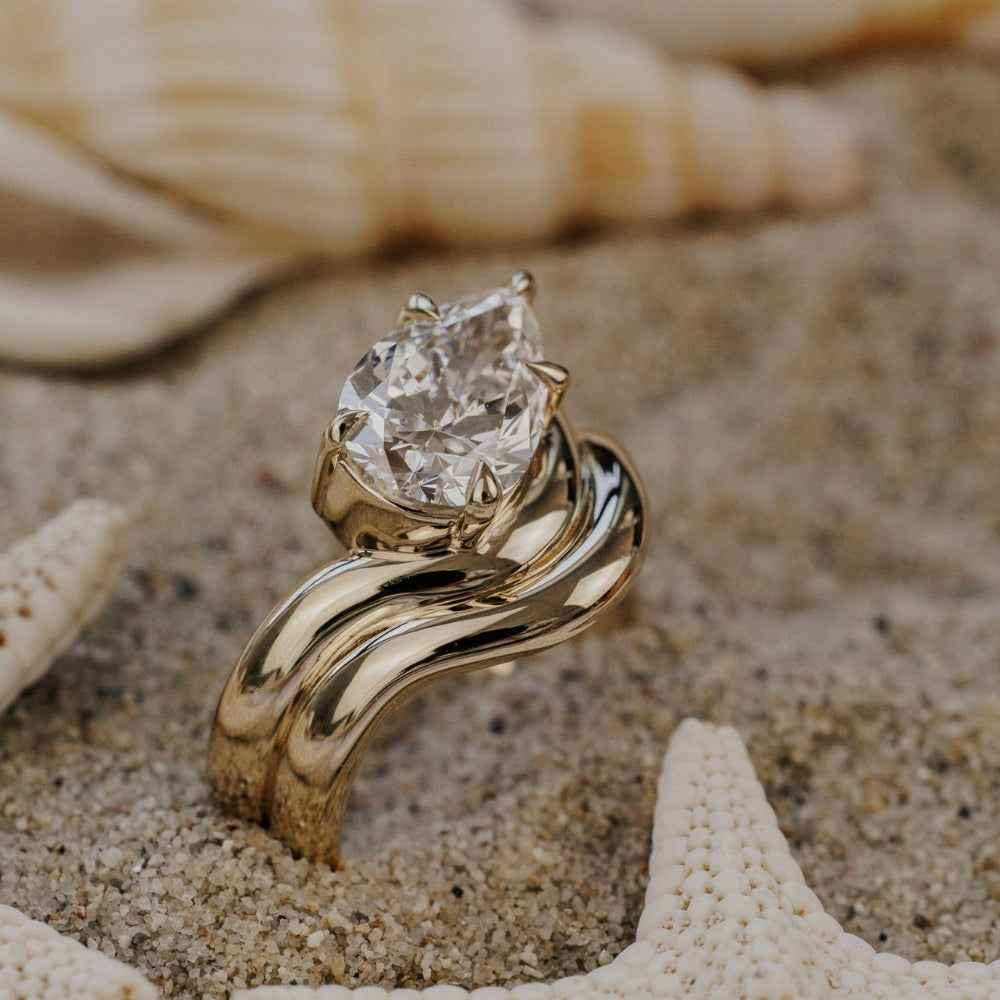 The Capri Pear Ring in 14k yellow gold featuring a lab-grown pear diamond prong set on a double wavy band, displayed in sand alongside a starfish.