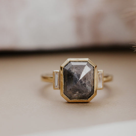 The Carson 5.71 ct Emerald Cut Rustic Diamond Ring in 14k yellow gold featuring an emerald cut gray and black rustic diamond in a milgrain bezel with baguette diamond side stones, displayed on a cream surface. This 14k yellow gold ring features a one-of-a-kind 5.71 ct emerald cut gray and black rustic natural diamond with .13 cttw baguette natural diamond side stones, all set in a milgrain bezel design.