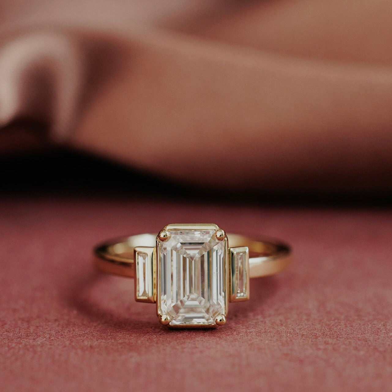 The Carson Emerald Cut Ring in 14k yellow gold featuring an emerald cut center stone with baguette diamond side stones, displayed on a dusty rose surface. This 14k gold ring features an emerald cut center stone set in four prongs with two ~0.13 cttw baguette natural diamond side stones. The Carson Emerald Cut Ring is made just for you based on the customizations you select. Make it yours by selecting your metal color, center stone type, and center stone size.
