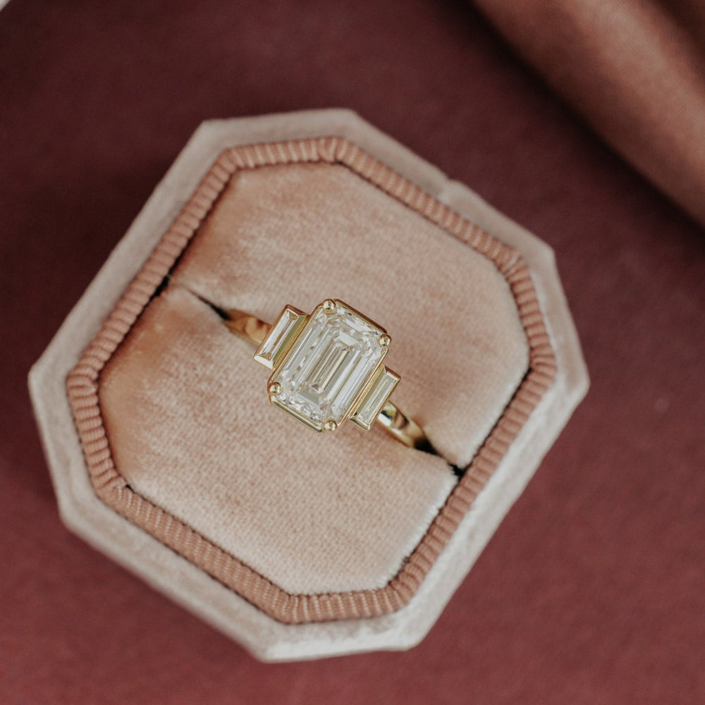 The Carson Emerald Cut Ring in 14k yellow gold featuring an emerald cut center stone with baguette diamond side stones, displayed in a blush velvet ring box. This 14k gold ring features an emerald cut center stone set in four prongs with two ~0.13 cttw baguette natural diamond side stones. The Carson Emerald Cut Ring is made just for you based on the customizations you select. Make it yours by selecting your metal color, center stone type, and center stone size.
