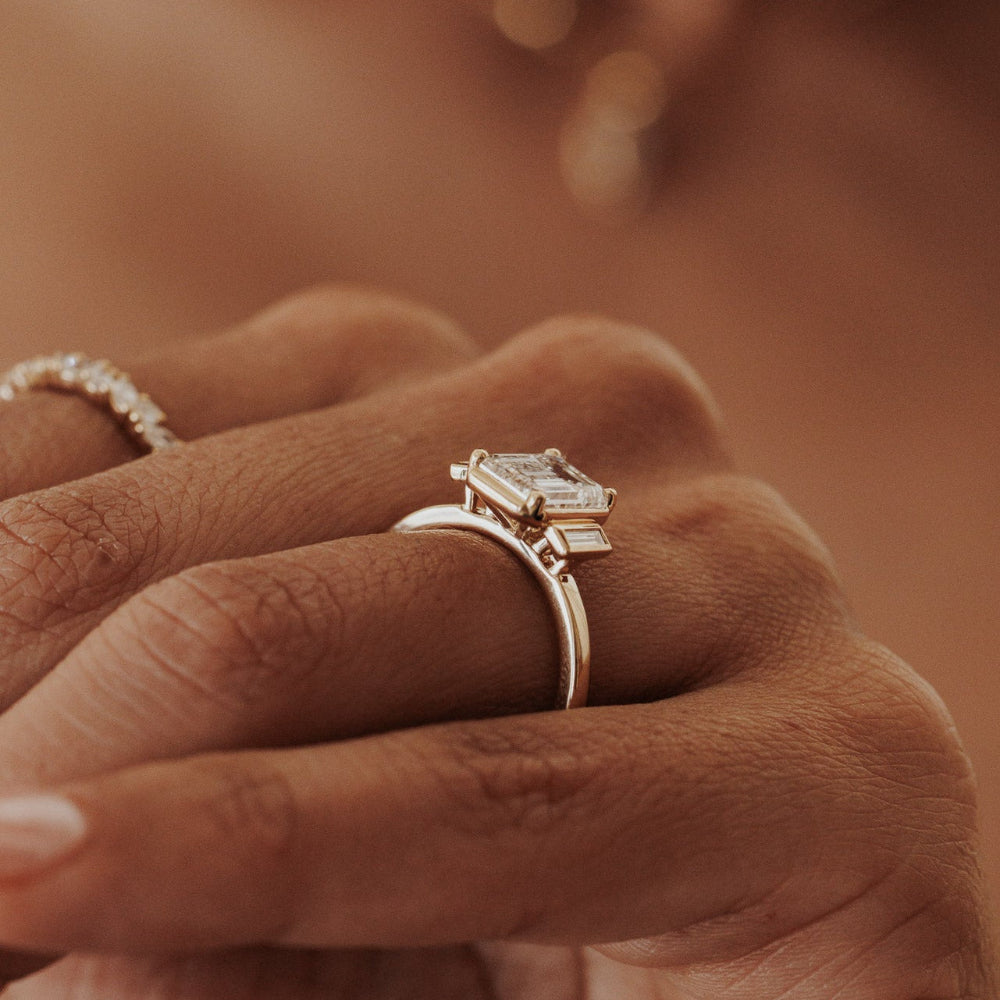 The Carson Emerald Cut Ring in 14k yellow gold featuring an emerald cut center stone with baguette diamond side stones, worn on the ring finger alongside a diamond eternity band on another finger.
