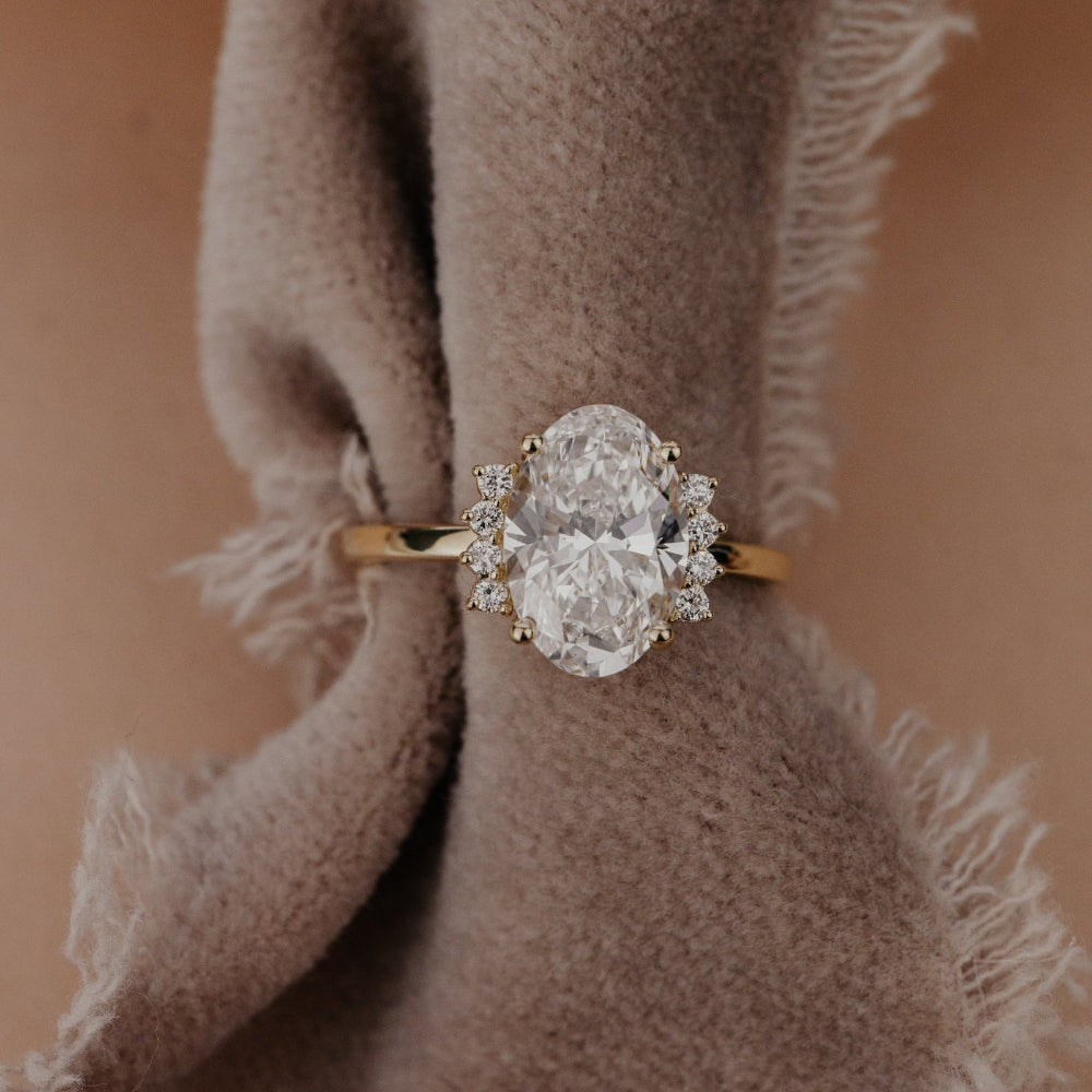 The Claire Oval Ring in 14k yellow gold featuring a lab grown diamond oval center stone set in four prongs, flanked by starburst-set accent diamonds on a high polish band, displayed on a soft textured fabric prop.