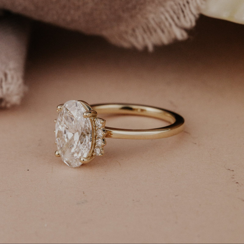The Claire Oval Ring in 14k yellow gold featuring a lab grown diamond oval center stone set in four prongs with starburst-set accent diamonds, shown from a side profile angle on a warm neutral surface with a soft fabric backdrop.