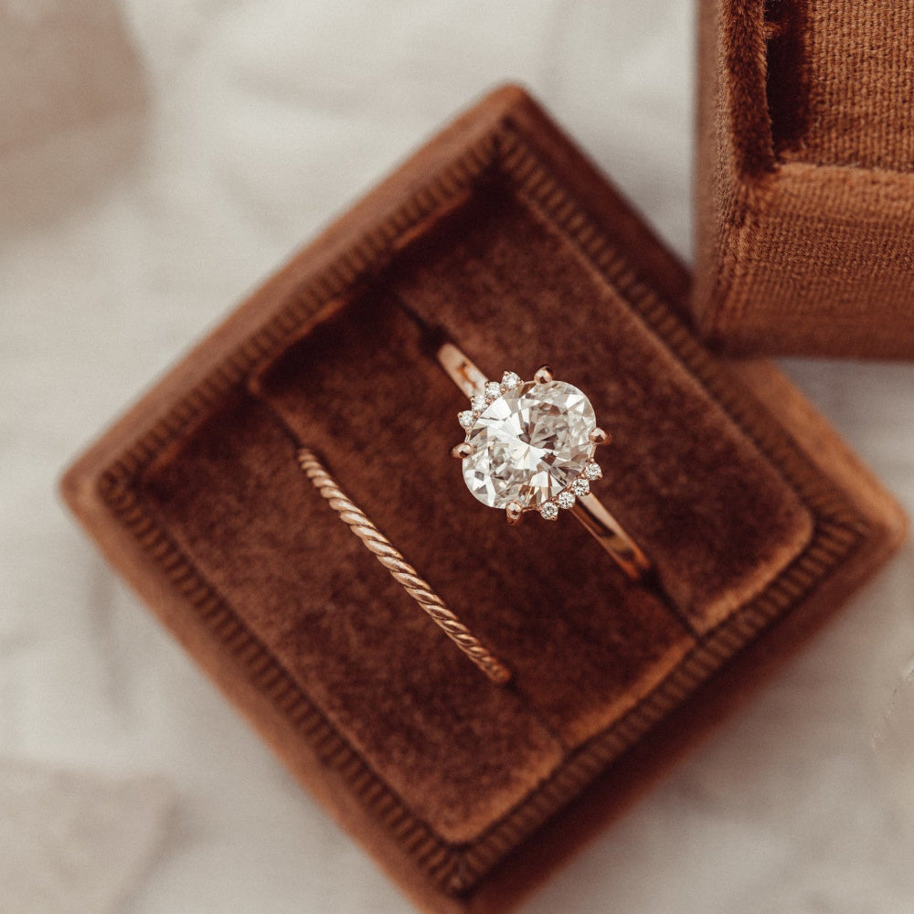 The Claire Oval Ring in 14k rose gold featuring a lab grown diamond oval center stone with starburst-set accent diamonds, displayed in a brown velvet ring box alongside a twisted stacking band.