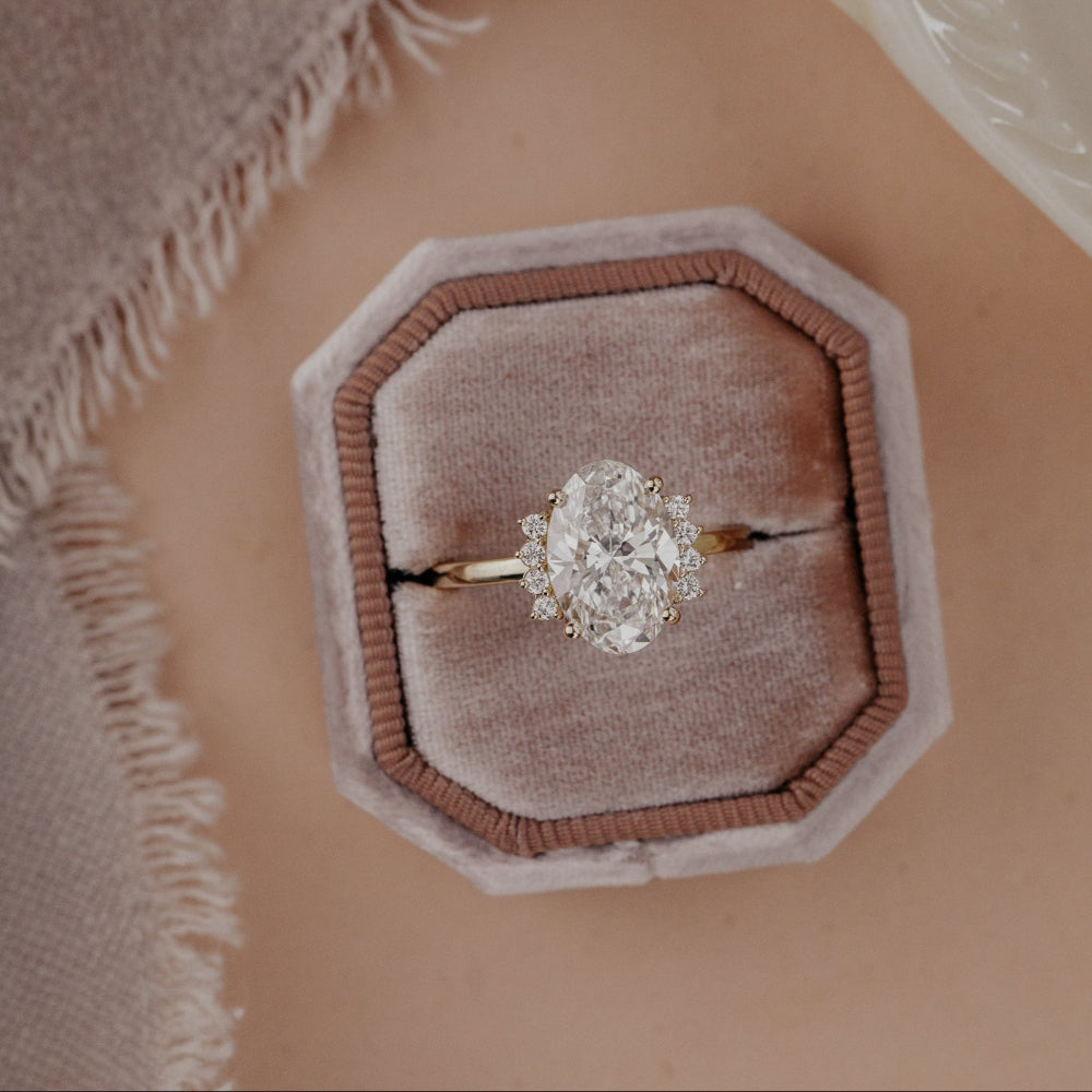 The Claire Oval Ring in 14k yellow gold with a lab grown diamond oval center stone and starburst-style side diamonds, displayed from above in a dusty pink octagonal velvet ring box.