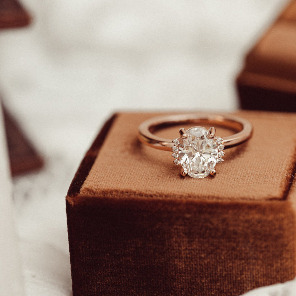 The Claire Oval Ring in 14k rose gold featuring a lab grown diamond oval center stone with starburst-set accent diamonds, displayed upright on the edge of a brown velvet ring box.