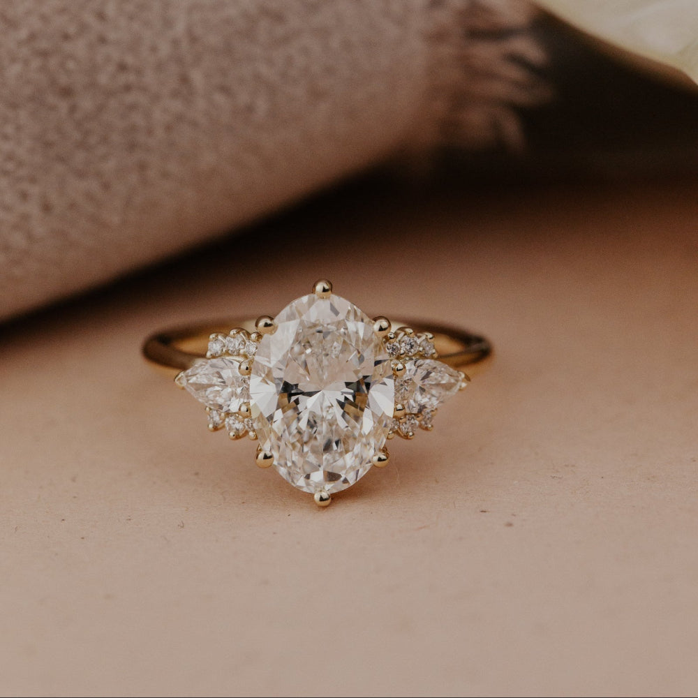 The Daphne Oval Ring in 14k yellow gold featuring a lab grown diamond oval center stone set in six prongs, flanked by pear and round diamond clusters on a high polish band.