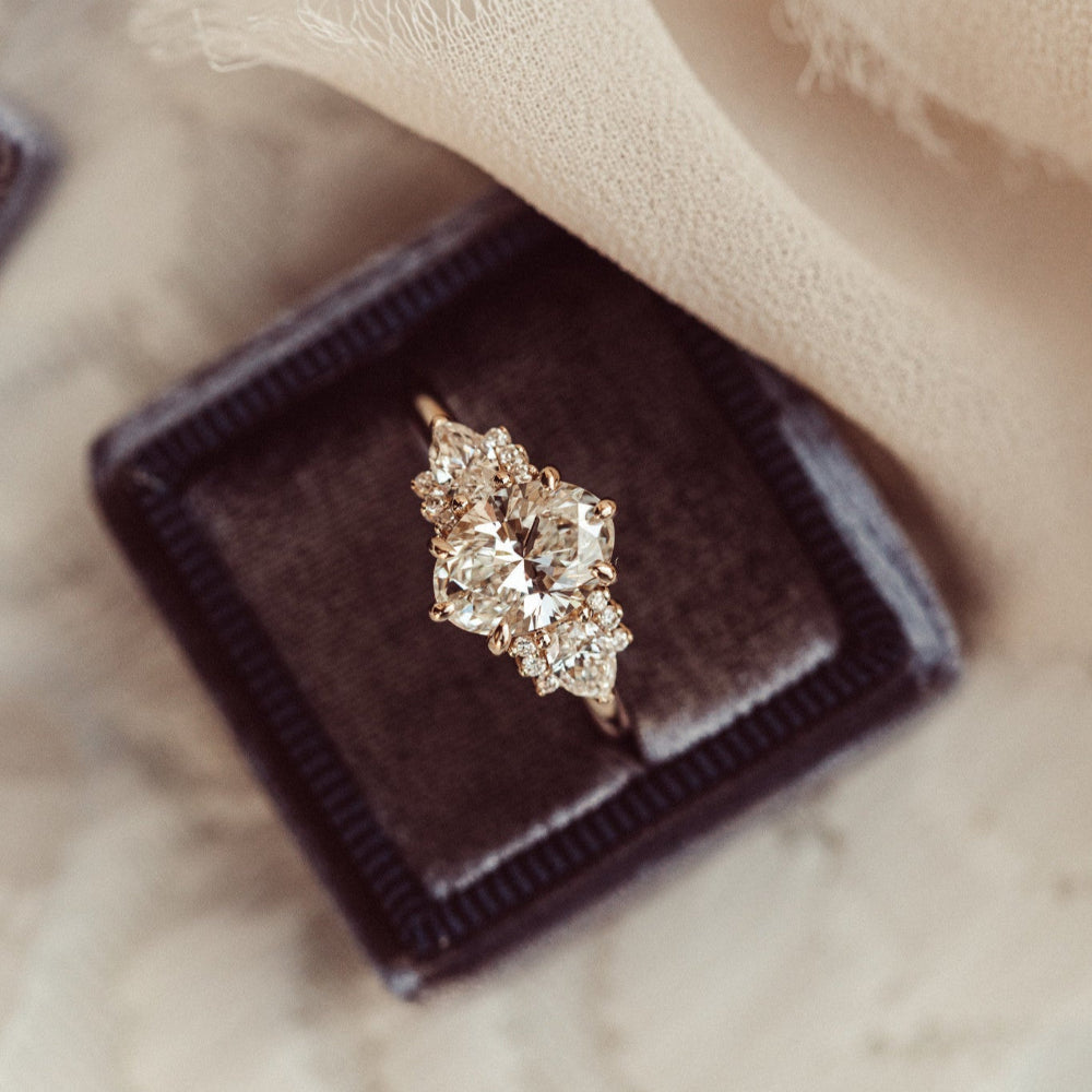 The Daphne Oval Ring in 14k rose gold featuring a lab grown diamond oval center stone set in six prongs with pear and round diamond clusters on a high polish band, displayed from above in a dark charcoal velvet ring box.