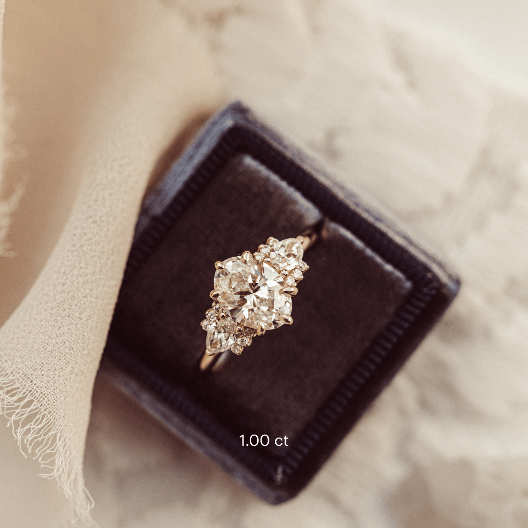 The Daphne Oval Ring in 14k rose gold featuring a lab grown diamond oval center stone set in six prongs with pear and round diamond clusters on a high polish band, displayed from above in a dark charcoal velvet ring box.