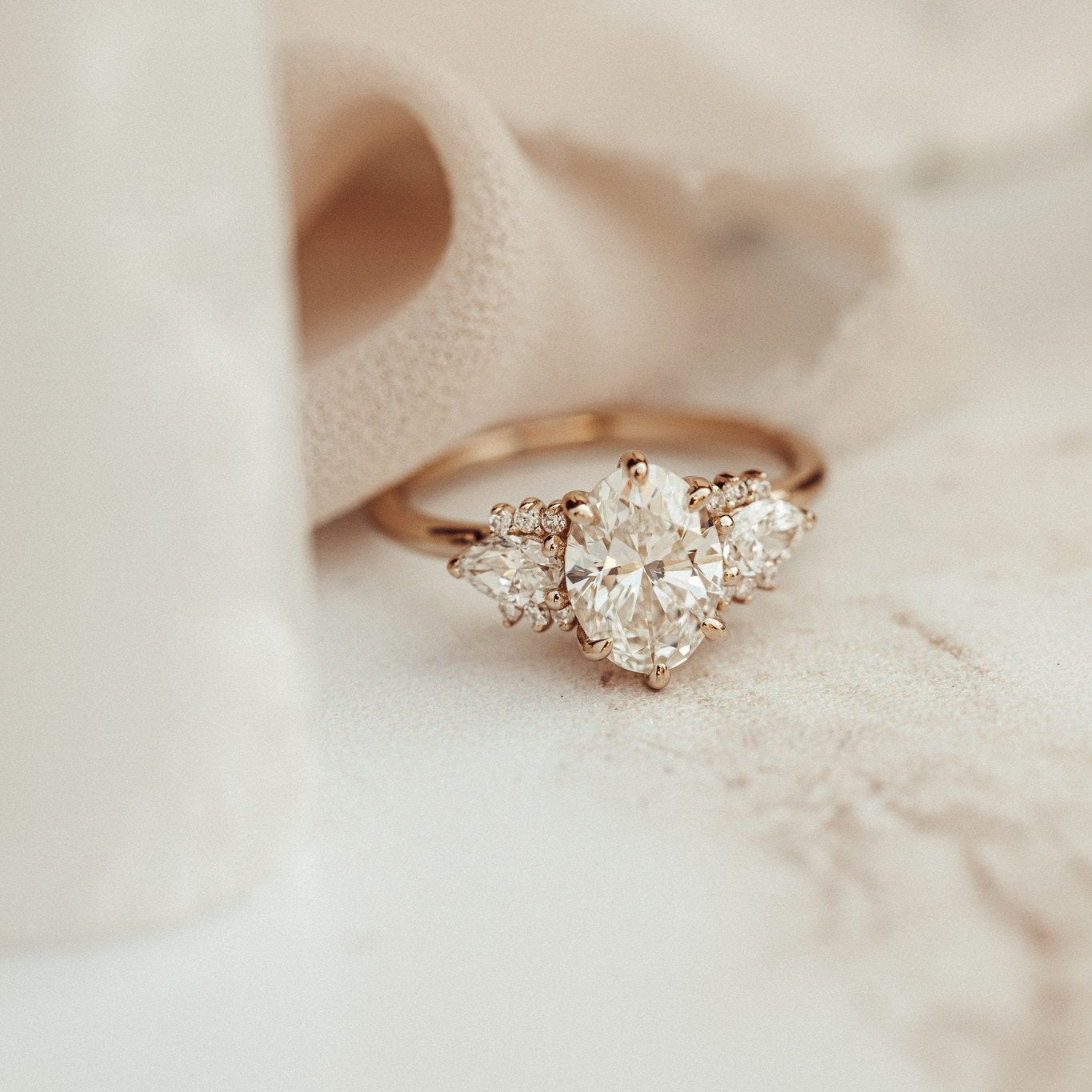 The Daphne Oval Ring in 14k rose gold featuring a lab grown diamond oval center stone set in six prongs with pear and round diamond clusters on a high polish band, styled on a soft textured surface.
