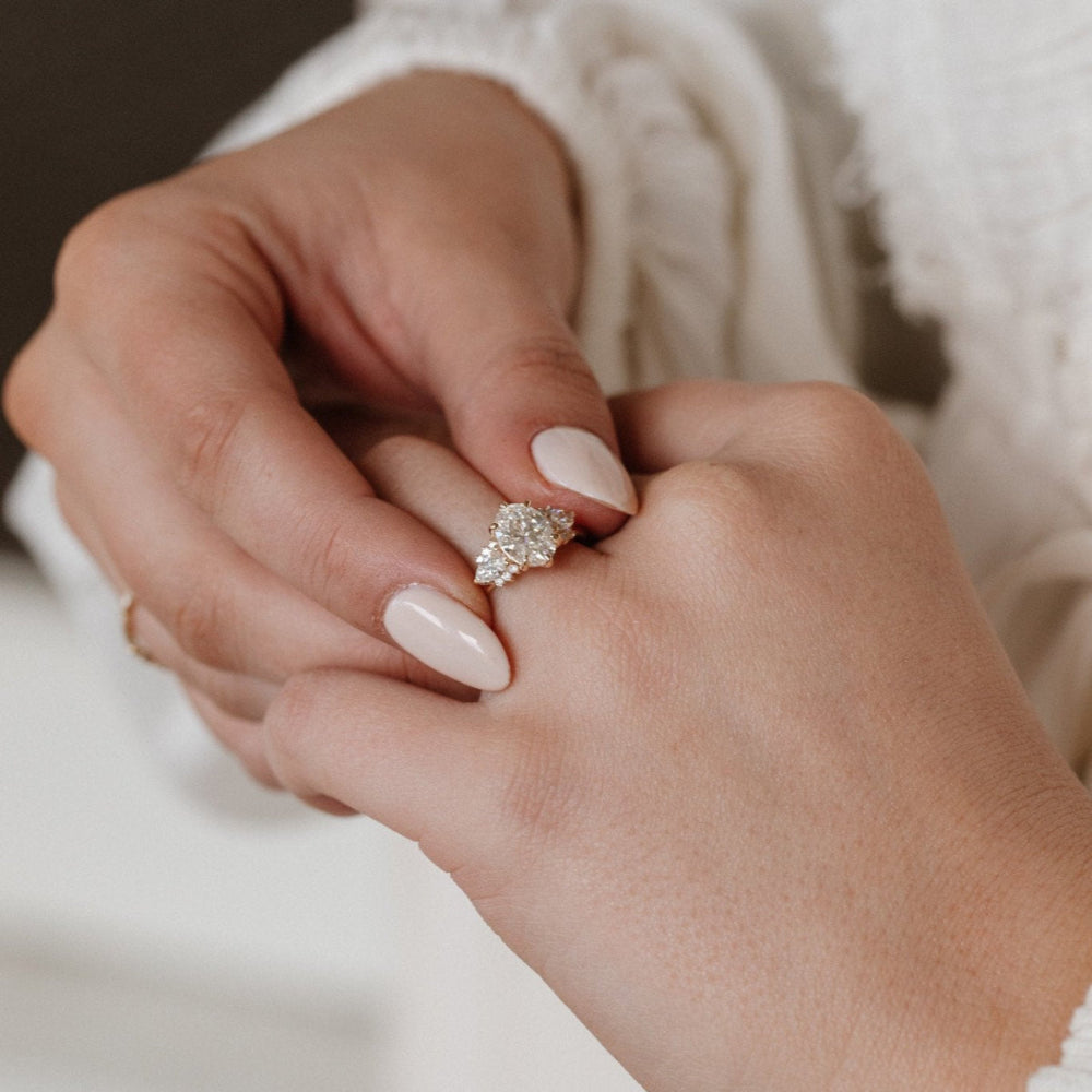 The Daphne Oval Ring in 14k yellow gold with a lab grown diamond oval center stone and pear and round diamond clusters, worn on a hand against a white textured background.