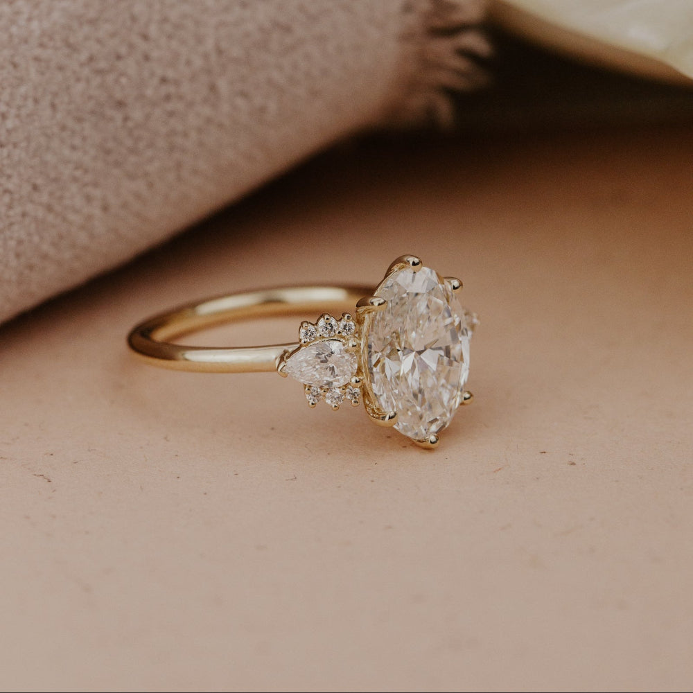 The Daphne Oval Ring in 14k yellow gold featuring a lab grown diamond oval center stone with pear and round diamond clusters, shown from a side profile angle on a warm neutral surface with a soft fabric backdrop.