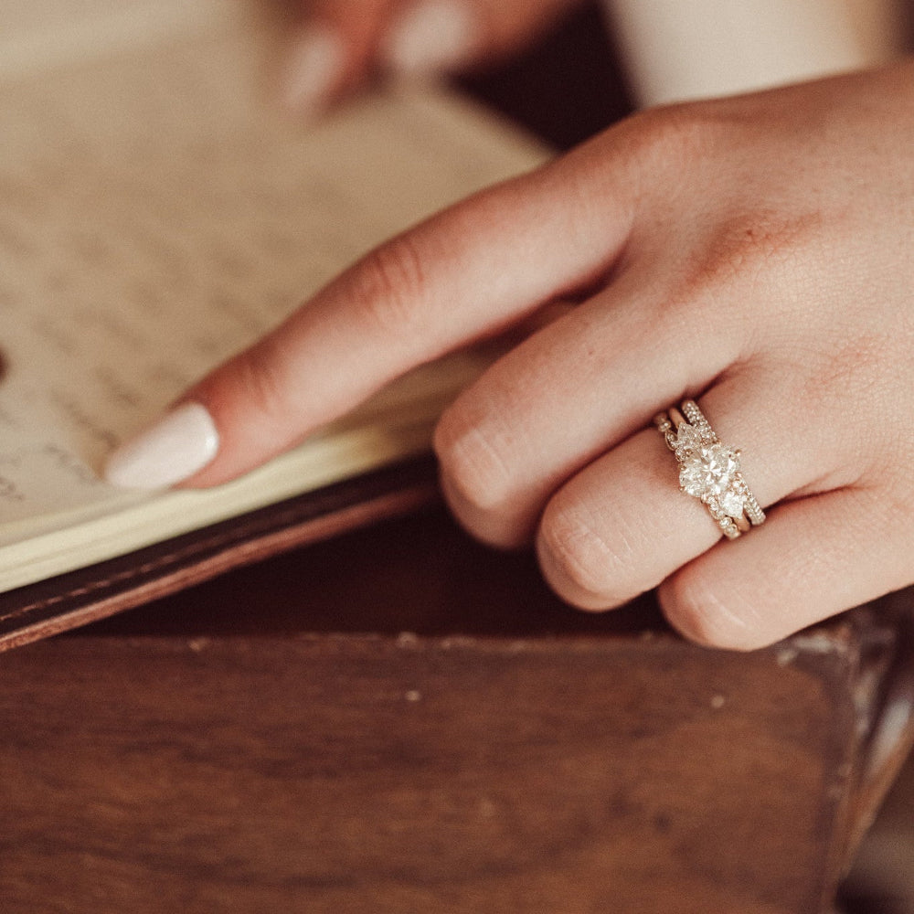 The Daphne Oval Ring in 14k yellow gold with a lab grown diamond oval center stone and pear and round diamond clusters, worn stacked with a wedding band on a model's hand as she writes in a journal.