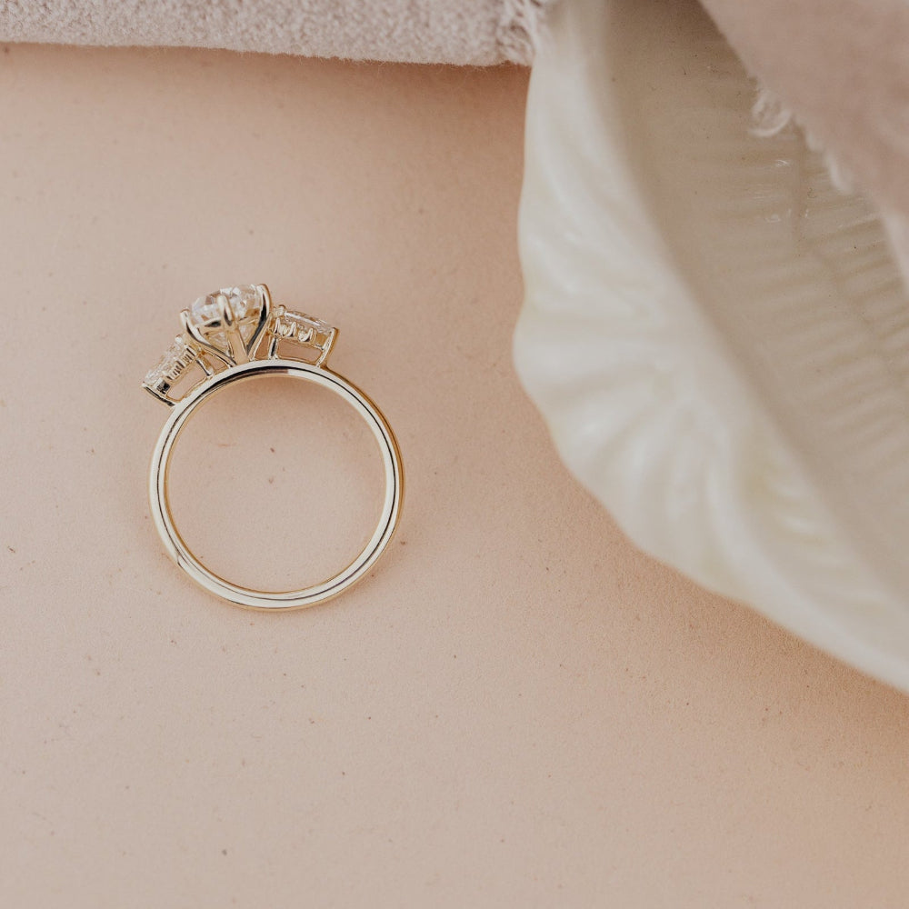 The Daphne Oval Ring in 14k yellow gold featuring a lab grown diamond oval center stone with pear and round diamond clusters, shown standing upright from an overhead angle on a warm neutral surface.