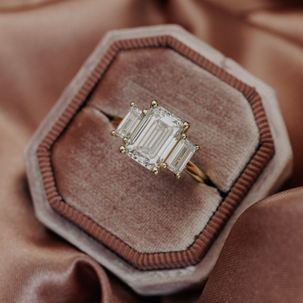 The Hannah Emerald Cut Ring in 14k yellow gold, featuring an emerald cut center stone set in four prongs with two large emerald cut diamond side stones, displayed in an octagonal velvet ring box.