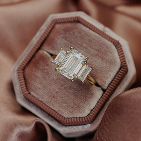 The Hannah Emerald Cut Ring in 14k yellow gold, featuring an emerald cut center stone set in four prongs with two large emerald cut diamond side stones, displayed in an octagonal velvet ring box.