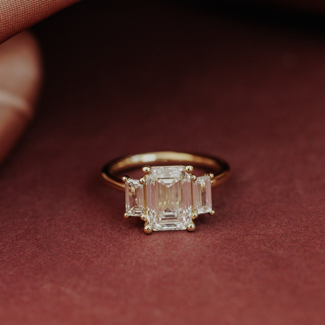The Hannah Emerald Cut Ring in 14k yellow gold, featuring an emerald cut center stone set in four prongs with two large emerald cut diamond side stones, shown from a front-facing angle on a deep red surface.

