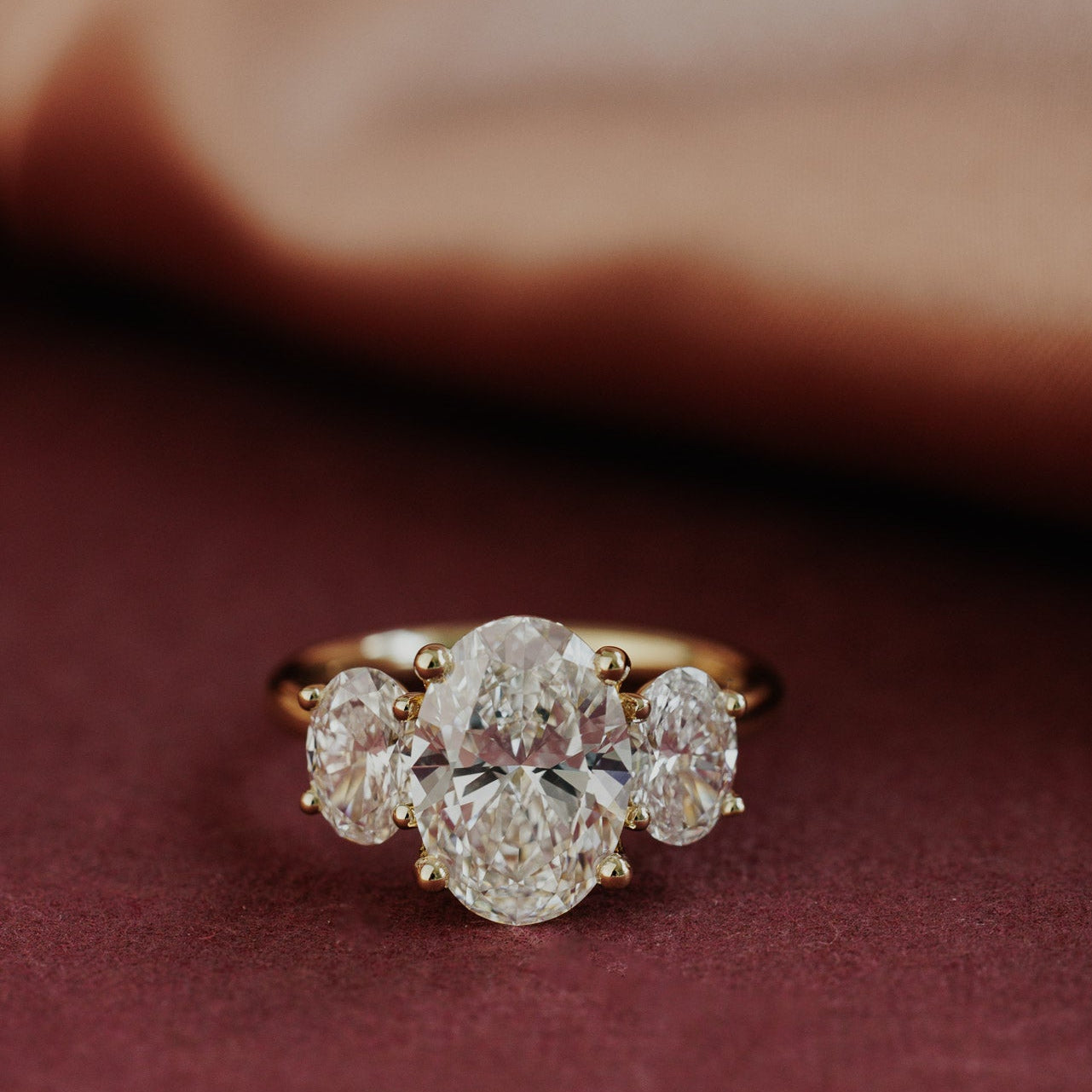 The Hannah Oval Ring in 14k yellow gold, featuring an oval cut center stone set in four prongs with two large oval diamond side stones, shown from a front-facing angle on a deep red surface.
