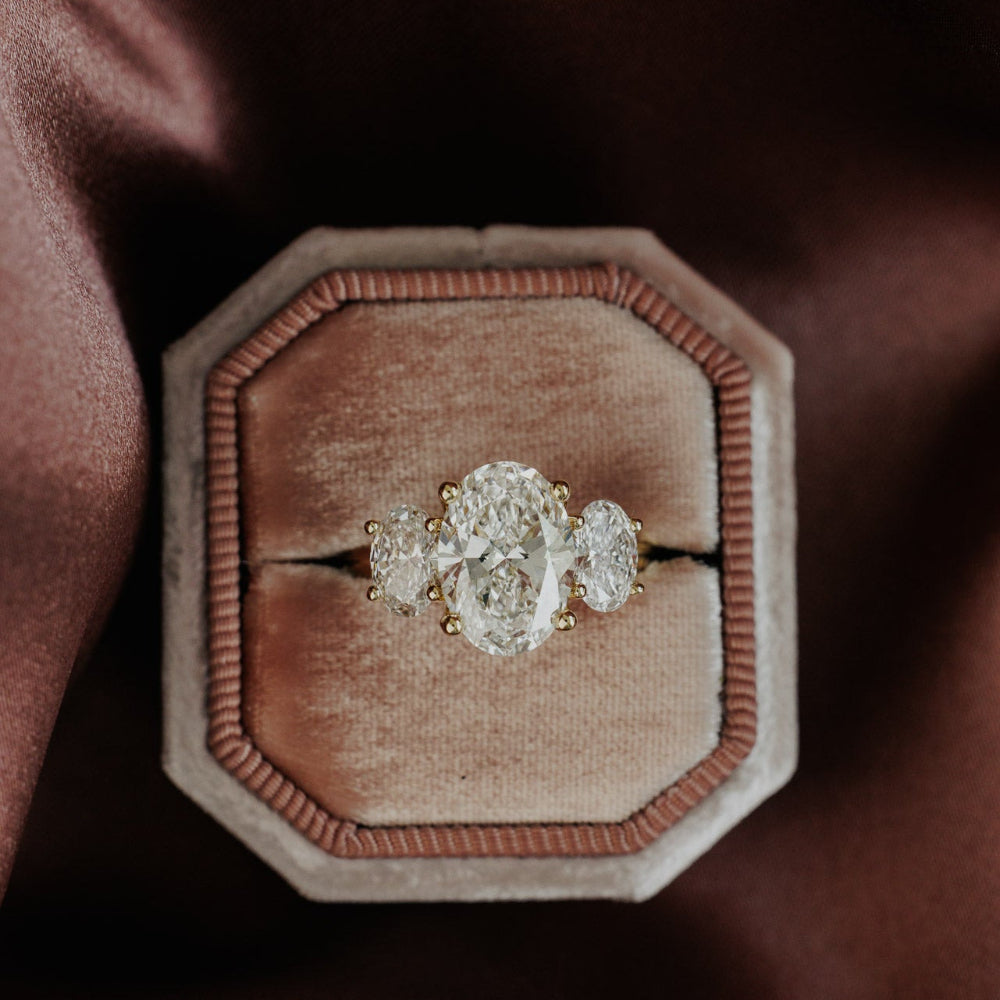 The Hannah Oval Ring in 14k yellow gold, featuring an oval cut center stone set in four prongs with two large oval diamond side stones, displayed in an octagonal velvet ring box.
