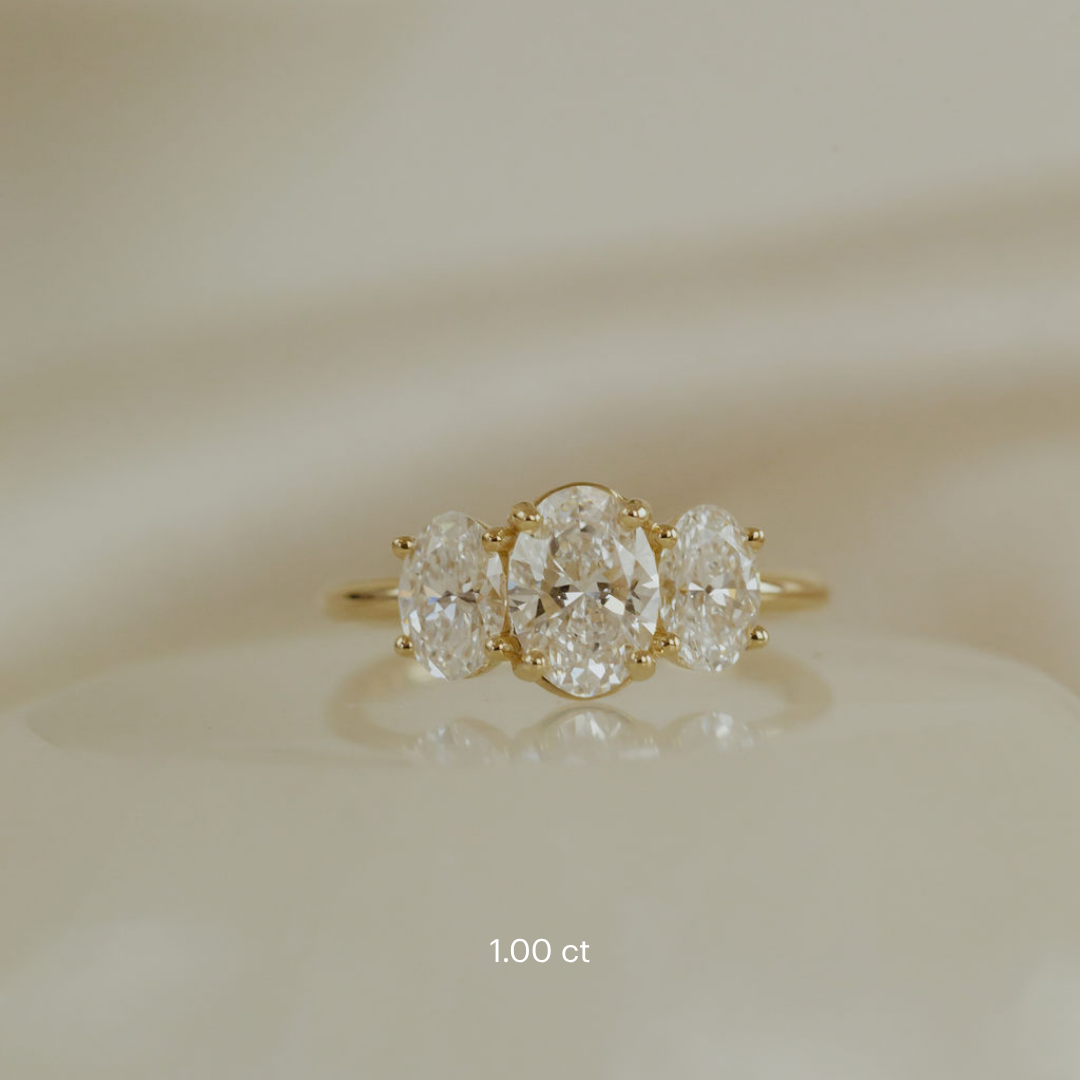 The Hannah Oval Ring in 14k yellow gold, featuring an oval cut center stone set in four prongs with two large oval diamond side stones, shown in the 1.00 ct center stone size on a cream surface.
