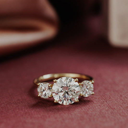 The Hannah Round Ring in 14k yellow gold, featuring a round cut center stone set in four prongs with two large round diamond side stones, shown from a front-facing angle on a deep red surface.