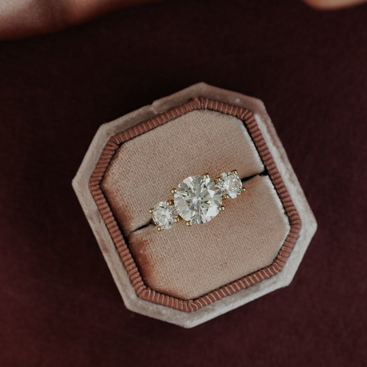The Hannah Round Ring in 14k yellow gold, featuring a round cut center stone set in four prongs with two large round diamond side stones, displayed in an octagonal velvet ring box.
