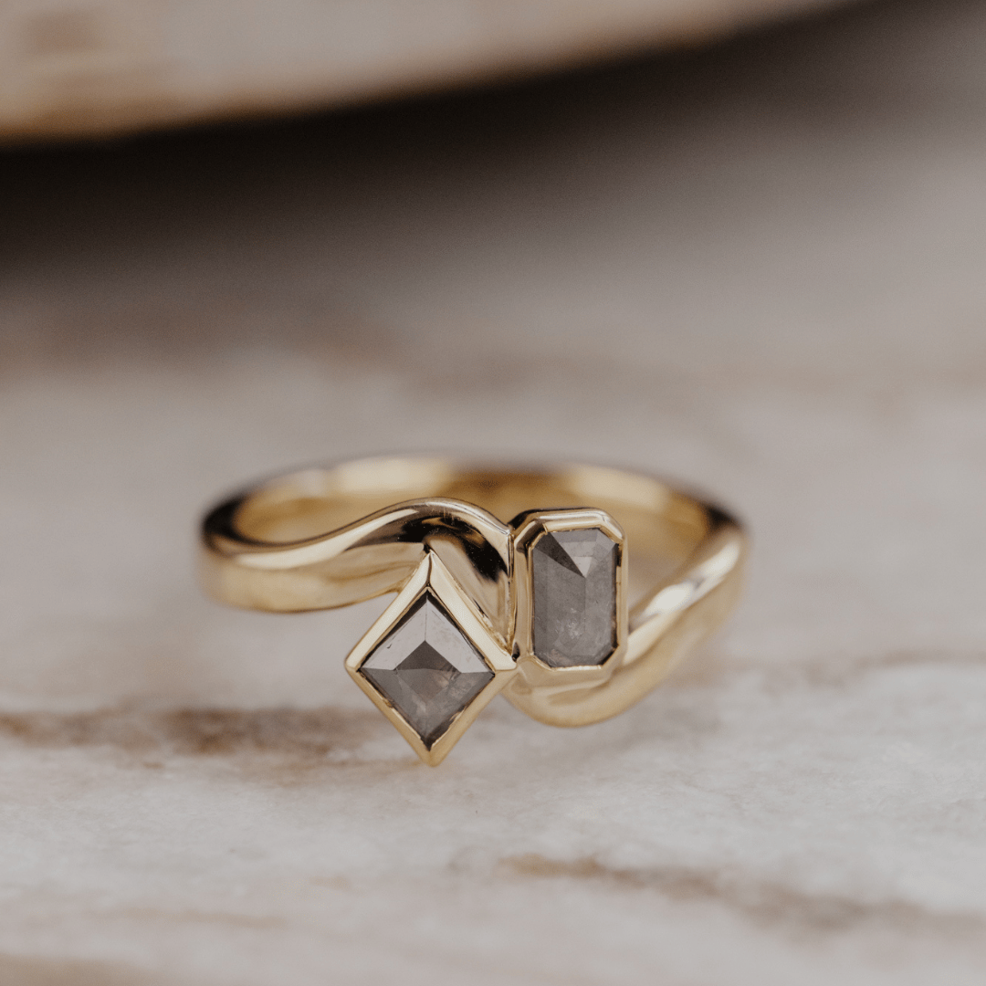 A gold ring with two gray rustic diamonds resting on a light marble surface.