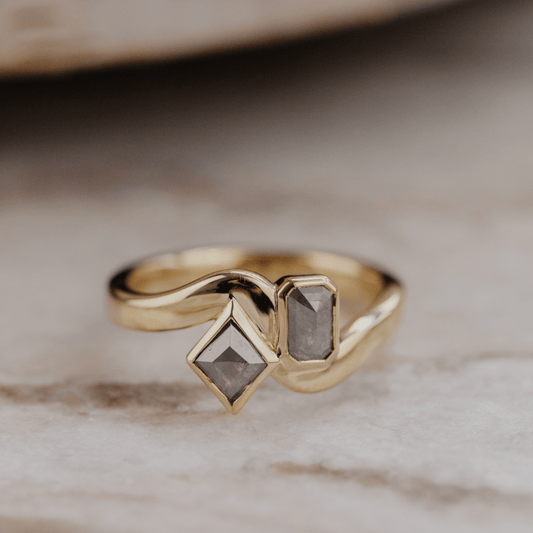 A gold ring with two gray rustic diamonds resting on a light marble surface.