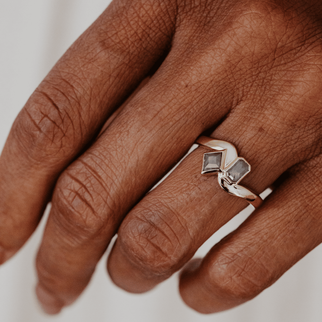 A close-up of a hand wearing a yellow gold ring with two gray rustic diamonds set against a soft, light gray background.