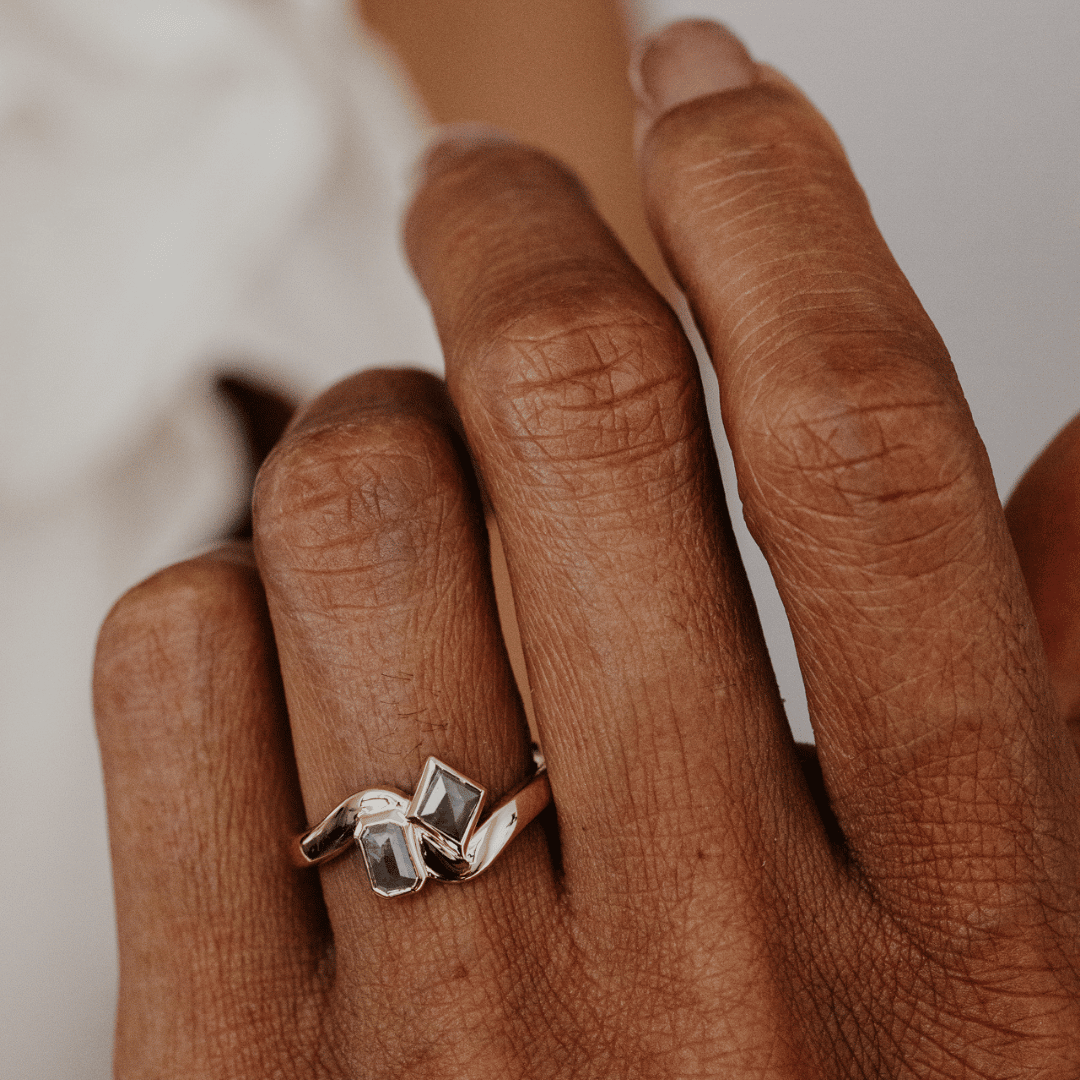A close-up of a person's hand wearing a gold ring with three rectangular clear stones set in a wavy, intertwined design.