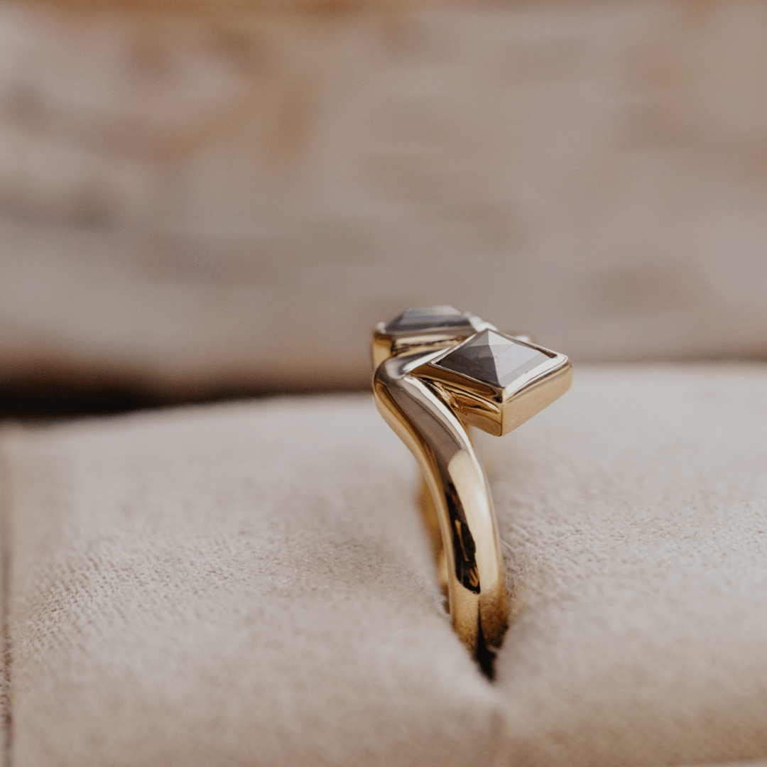 A gold ring with a unique sculptural band and a square-cut gray rustic diamonds displayed on a textured beige ring box.