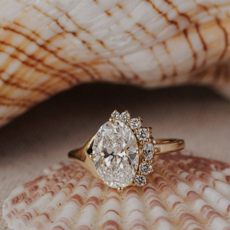 The Horizon Oval Ring in 14k yellow gold featuring a lab-grown oval diamond center stone with a round tapered diamond half halo, displayed inside a seashell. This 14k gold ring features an oval center stone, surrounded by a ~.22 cttw round tapered diamond half halo. The entire design is set flush to the finger. This ring is made-to-order based on the selected customizations.