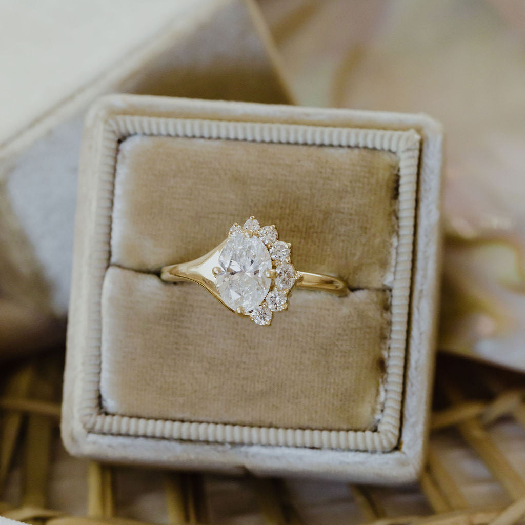 The Horizon Oval Ring in 14k yellow gold featuring a lab-grown oval diamond center stone with a round tapered diamond half halo, displayed in a tan velvet ring box. This 14k gold ring features an oval center stone, surrounded by a ~.22 cttw round tapered diamond half halo. The entire design is set flush to the finger. This ring is made-to-order based on the selected customizations.
