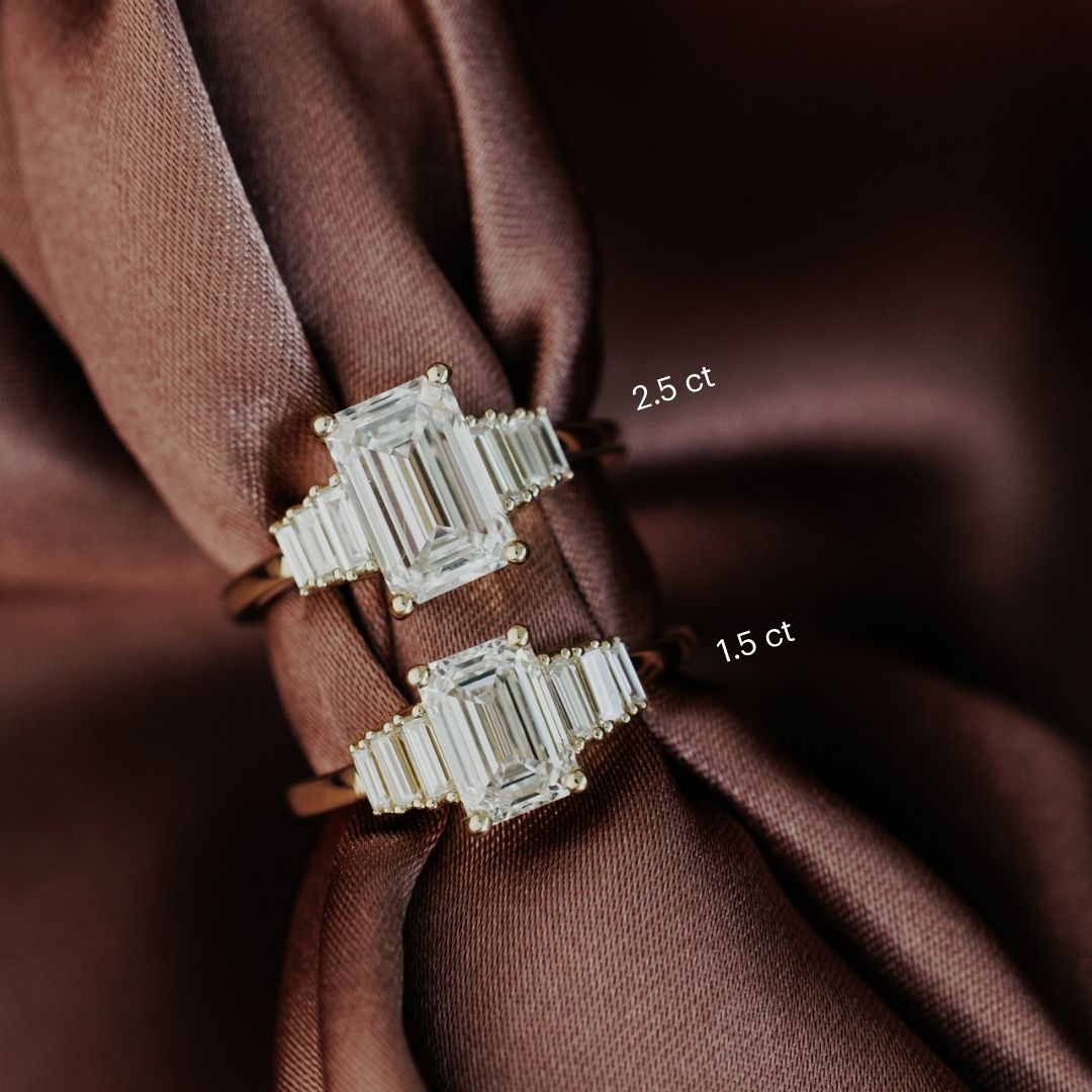 The Jolee Emerald Cut Ring in 14k yellow gold shown in two carat weight options: 2.5 ct (top) and 1.5 ct (bottom), displayed on draped brown satin fabric.
