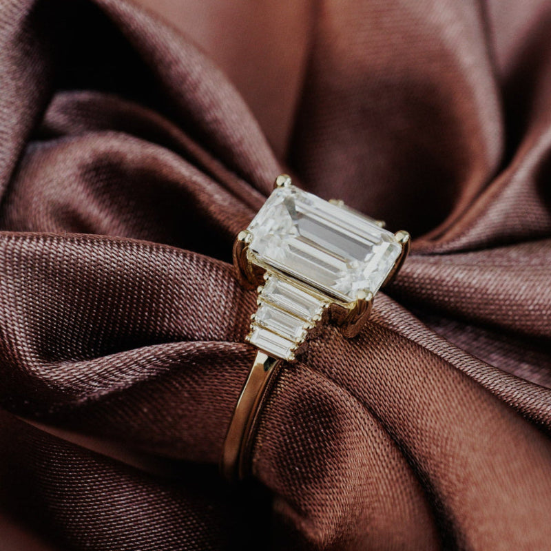 The Jolee Emerald Cut Ring in 14k yellow gold, featuring an emerald cut center stone set in four prongs with baguette natural diamond side stones, shown from a front-facing angle on draped brown satin fabric.
